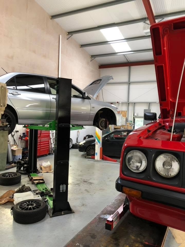 A car raised on a two-post lift for repairs, beside a classic with its bonnet up